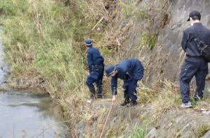 警察官が川岸の藪で行方不明者を捜索している写真です。